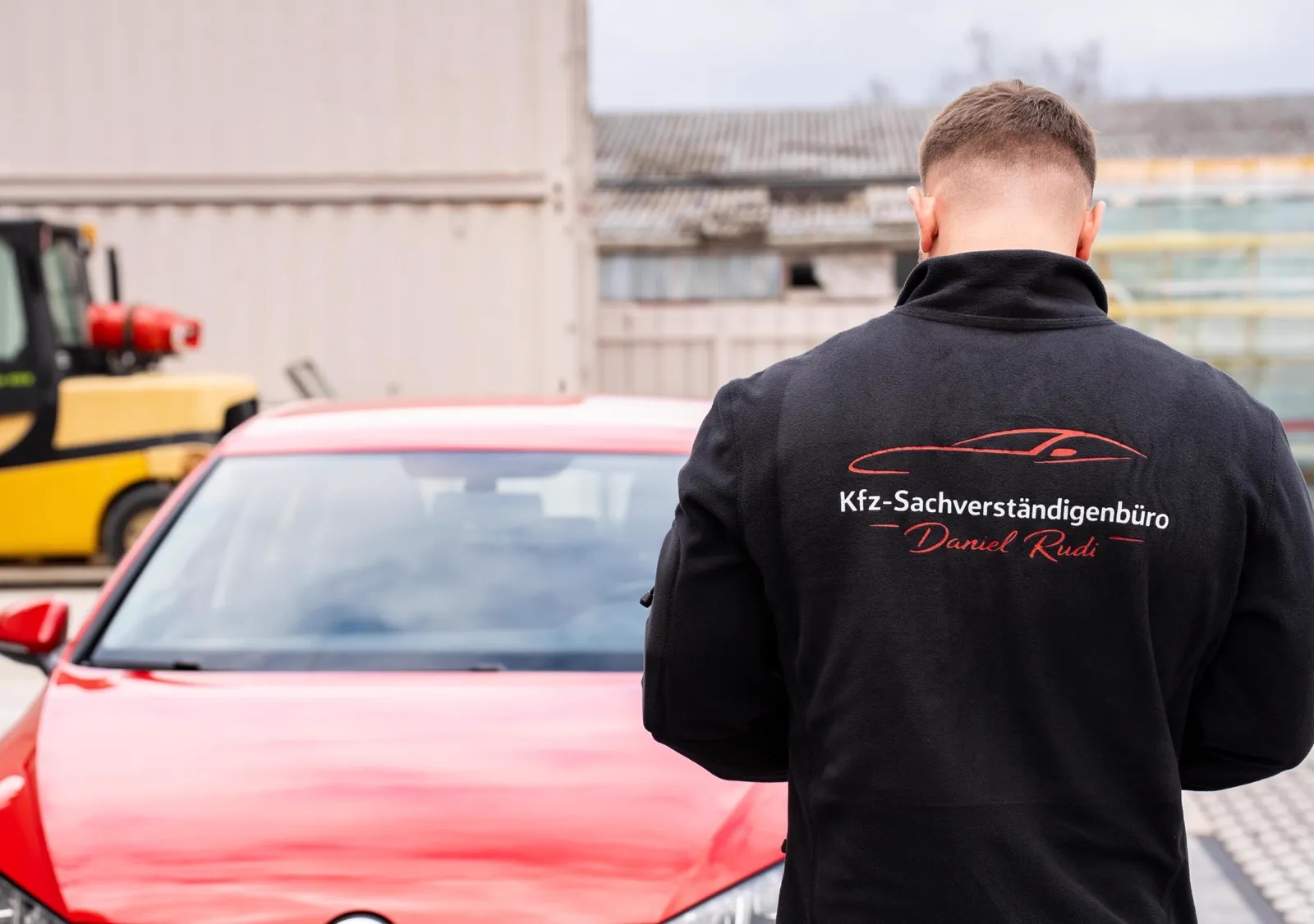 Kfz-Sachverständiger Daniel Rudi in Arbeitsjacke mit Firmenlogo vor einem roten Fahrzeug bei der Bewertung